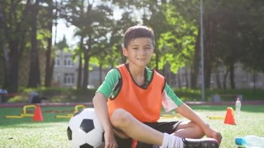 Turuncu yelekli yakışıklı Asyalı çocuk futbol topuyla yapay sahada oturuyor ve kameraya bakıyor.