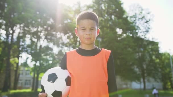 Futbolista de 13 años cansado en chaleco naranja sosteniendo la pelota en su mano y mirando a la cámara en el campo de deportes al aire libre en un día soleado — Vídeos de Stock