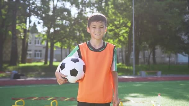 Bello stanco asiatico teen ragazzo in calcio abbigliamento in piedi con palla su il stadio tra città parco alberi e guardando fotocamera con grave faccia — Video Stock