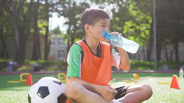 Bonito cansado desportivo 13-aged futebolista sentado na cobertura de grama especial do terreno esporte ao ar livre e água de dringking após o treinamento no dia ensolarado — Vídeo de Stock