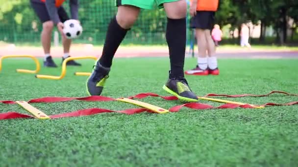 Vista frontal de los pies de los niños en zapatos de fútbol que hacen diferentes ejercicios de fútbol en la escalera durante el día de entrenamiento en el campo de deporte al aire libre — Vídeos de Stock