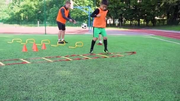 Voetbal trainingsdag waar een tienerjongen joggen op de voetballadder terwijl een andere speler traint de kick samen met professionele trainer op het voetbalveld — Stockvideo