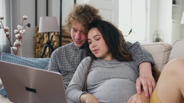 Bonito amor sorridente jovem beijando na testa sua encantadora esposa grávida feliz com dreadlocks enquanto assiste vídeo interessante no laptop em casa — Vídeo de Stock