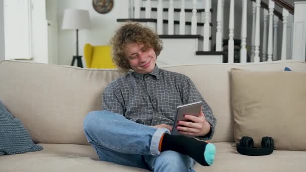 Gros plan de beau sourire marié de 30 ans avec les cheveux bouclés qui sont assis sur le canapé doux dans le salon et en utilisant i-pad pour voir la vidéo intéressante — Video