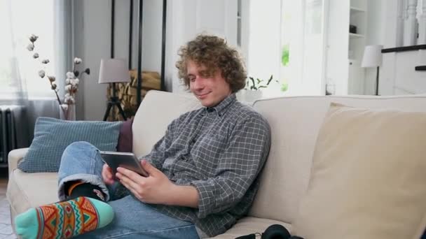 Hombre casado joven satisfecho de buen carácter con el pelo rizado vestido con clavos casuales sentado en el acogedor sofá en el apartamento contemporáneo y el uso de la tableta pc — Vídeo de stock