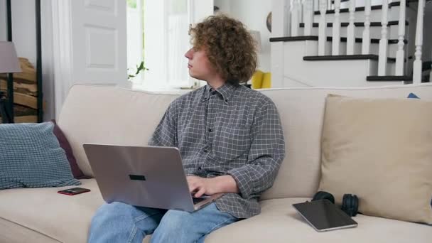 Pensativo confiante criativo jovem casado homem sentado no sofá na sala de estar e trabalhando no laptop simultaneamente olhando para a janela — Vídeo de Stock