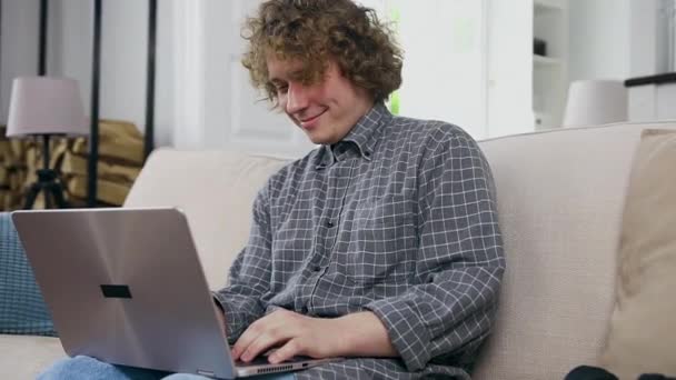 Sympathisch positiv lächelnder junger Kerl mit lockigem Haar, der zu Hause auf dem gemütlichen Sofa sitzt und lustige Videos auf dem Laptop anschaut, Frontansicht — Stockvideo