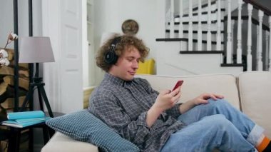 Memnuniyet verici kulaklıklı genç adam evdeki rahat kanepede telefon çalma listesinden müzik dinliyor.