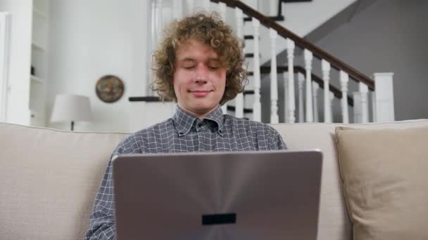 Przystojny pozytywny twórczy kręcone młody facet siedzi na wygodne kanapa w domu i pracuje na laptopie, widok z przodu — Wideo stockowe