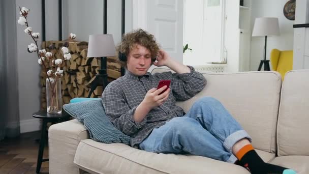 Красивий задоволений позитивний молодий хлопець відпочиває на зручному дивані у вітальні і насолоджується сучасною музикою в навушниках — стокове відео