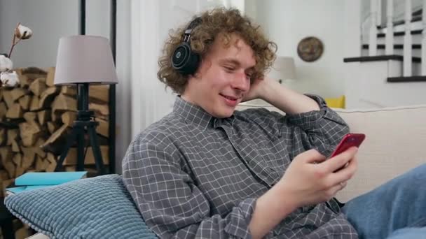 헤드폰을 낀 곱슬머리의 행복 한 남자가 집의 부드러운 소파에 앉아 모바일 플레이 리스트에서 음악을 듣고 있다. — 비디오