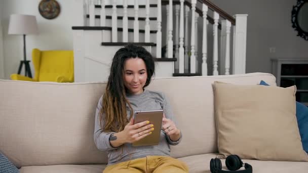 Обаятельная улыбающаяся стильная молодая беременная женщина, сидящая на удобном диване в гостиной и просматривающая любимые приложения на i-pad — стоковое видео