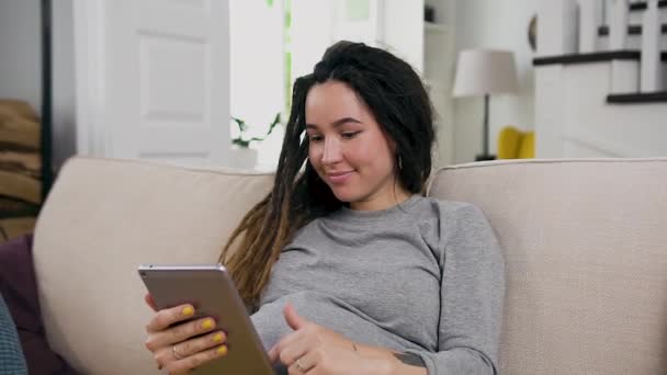 Bastante sonriente joven embarazada feliz con rastas relajarse en el sofá suave en casa y fotos de navegación en i-pad — Vídeo de stock