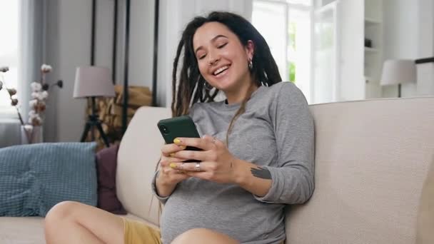 Utmärkt glad gift glad ung kvinna tittar roliga bilder på sin telefon och försiktigt röra hennes gravida mage — Stockvideo