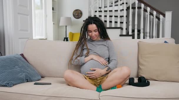 Будущая концепция материнства, где очаровательная счастливая, улыбающаяся замужняя девушка с дредами нежно трогает свой беременный живот и смотрит в камеру дома — стоковое видео