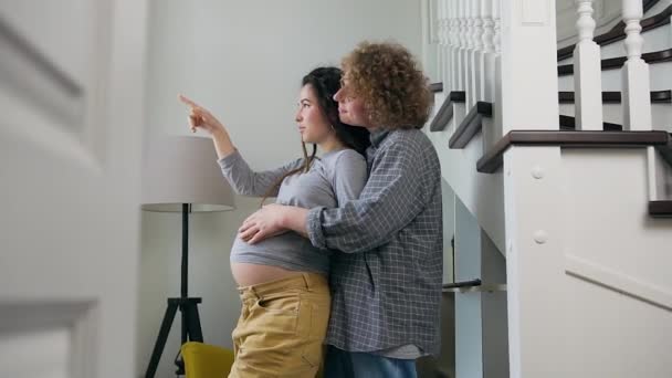 Beautiful happy trendy pregnant woman standing together with her loving curly husband in front of the window and showing something behind it outdoors — Stock Video