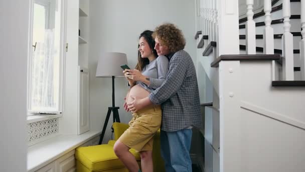 窓の前に立って将来の親を楽しむ魅力的な幸せな笑顔の若い夫婦妊娠中のカップル — ストック動画