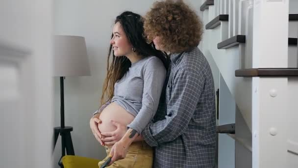 Gut aussehendes, fröhlich lächelndes junges schwangeres Paar genießt zukünftige Elternschaft, während es gemeinsam vor dem Fenster steht — Stockvideo