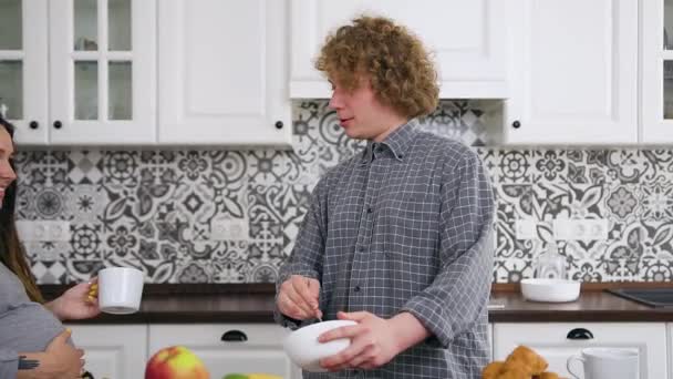 Hermosa mujer embarazada sonriente feliz con rastas sentado cerca de la mesa de la cocina, beber té y ver cómo su marido rizado preparar el desayuno — Vídeos de Stock