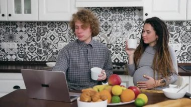 Modern mutfakta çay içen ve aynı zamanda dizüstü bilgisayar kullanan kıvırcık saçlı ve hamile kadınlardan oluşan çekici, pozitif evli genç bir ailenin ön görüntüsü.