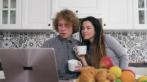 Прекрасний щасливий безтурботний молодий вагітна пара сидить біля кухонного столу, п'є чай і дивиться на екран ноутбука, вид спереду — стокове відео