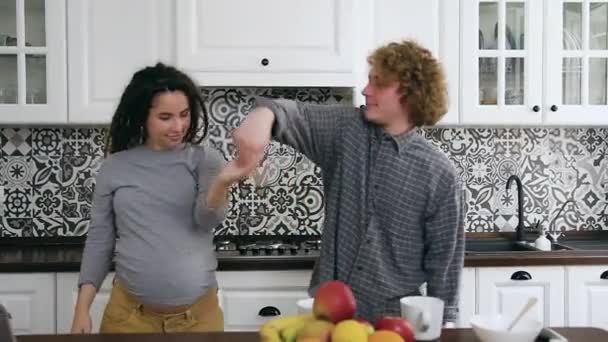 Front view of good-looking positive lucky young man and pregnant woman with dreadlocks which having fun together in the modern kitchen with dances, huggs and kisses — стоковое видео