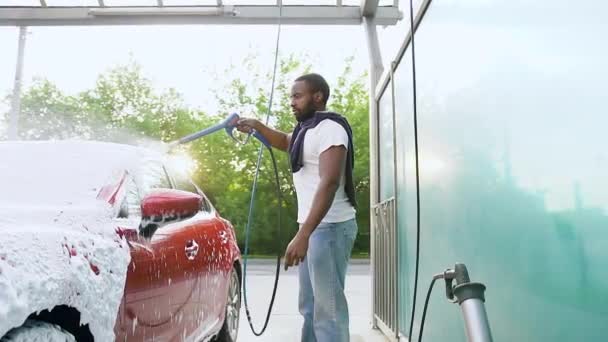 Provável grave jovem barbudo homem de pele preta lavando seu carro vermelho com tubo de jato com espuma seca em auto-serviço de lavagem de carro ao ar livre — Vídeo de Stock
