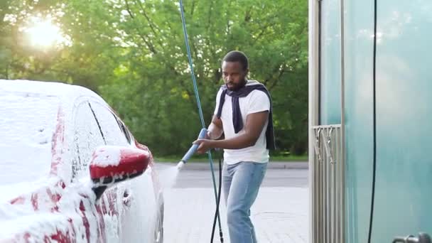 Attraente serio giovane ragazzo dalla pelle nera con barba lavare la sua auto costosa rossa all'aperto autolavaggio utilizzando tubo jet con schiuma di sapone — Video Stock