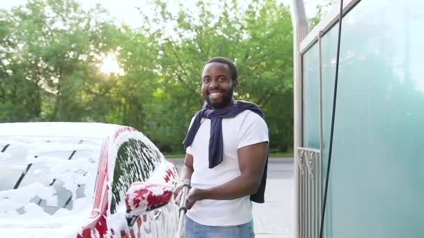 Bonito sorridente jovem barbudo americano africano de pé com tubo de jato com bico espumoso perto de seu carro em lavagem de carro moderno — Vídeo de Stock