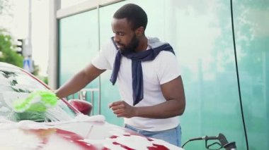 Yakışıklı, sakallı Afrikalı Amerikalı arabasını temizleme eldiveni ile yıkıyor.