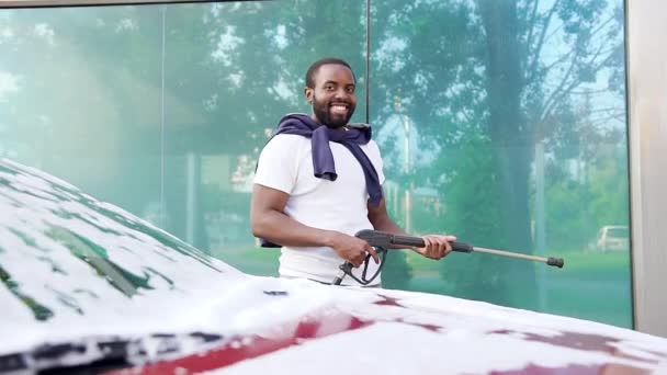 Close-up van positieve knappe jonge Afrikaanse Amerikaan die poseren op camera in de buurt van zijn luxe auto met hoge druk wasmachine in car wash — Stockvideo