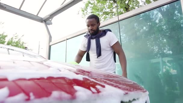 Gutmütig zufriedener junger bärtiger dunkelhäutiger Mann wäscht rotes Luxusauto mit Handschuhen und wischt Motorhaube aus Schaum in Waschanlage — Stockvideo