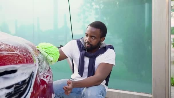 Όμορφος σίγουρος νεαρός Αφρικανός Αμερικανός σε πράσινο γάντι χειρός πλύσιμο προβολέων με αφρό στο πλυντήριο αυτοκινήτων self-service — Αρχείο Βίντεο