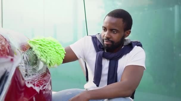 Attractive smiling happy bearded black-skinned guy looking at camera while washing his car with hand glove in car wash station — Stock Video