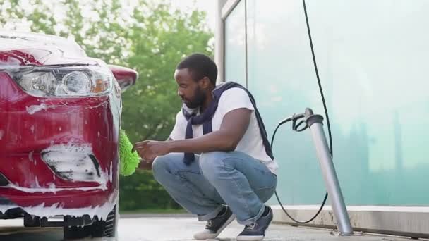 Красивый концентрированный бородатый молодой африканский американец чистит колеса своего автомобиля в ручных перчатках с пеной на автомойке — стоковое видео