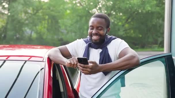 Attrayant moderne souriant barbu afro-américain debout près de sa voiture de luxe rouge et en utilisant le téléphone dans une journée ensoleillée parfaite — Video