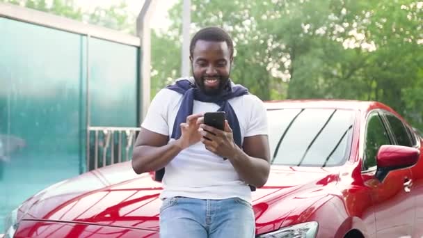 수염이 덥수룩하고 검은 피부의 젊은 남자가 자동차 세차장에서 자신의 붉은 비싼 차 옆에 서서 전화로 재구성 된 재밌는 사진을 즐기고 있습니다. — 비디오