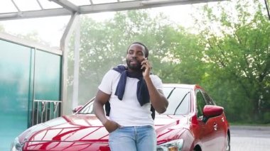 Çekici, genç, sakallı Afrikalı, sıradan giysiler içinde lüks bir arabanın yanında duruyor ve oto yıkamada telefonla konuşuyor.