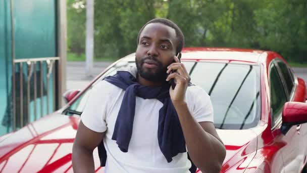 Vista frontal de la atractiva sonriente afroamericana de 30 años con barba bien arreglada en camiseta blanca que disfruta de su conversación telefónica cerca de su propio coche rojo brillante en el lavado de autos — Vídeos de Stock