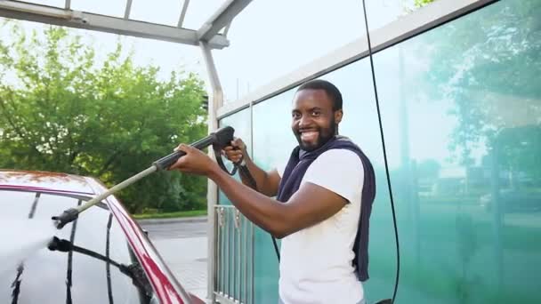 Primer plano de guapo sonriente joven barbudo afroamericano que mira a la cámara mientras lava su propio coche con alta presión de agua en el lavado de coches al aire libre — Vídeo de stock
