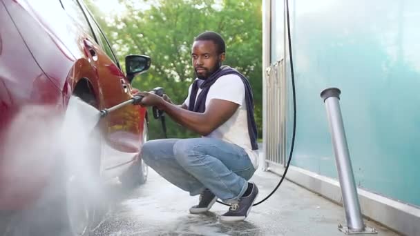 Vista frontal de simpático concentrado jovem barbudo homem de pele preta que lavar carros rodas usando máquina de lavar água de energia na lavagem de carro — Vídeo de Stock