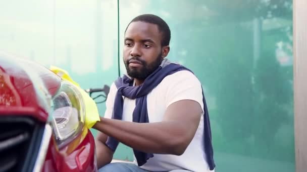 Sympatický mladý vousatý muž s černou pletí, který utírá světlomety žlutým ručníkem v myčce aut, zblízka — Stock video