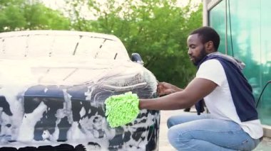 Yakışıklı, kendine güvenen, siyah sakallı, arabasının yanında çömelmiş, farlarını köpüklü süngerle yıkayan, açık hava oto yıkama alanında çalışan genç bir adam.