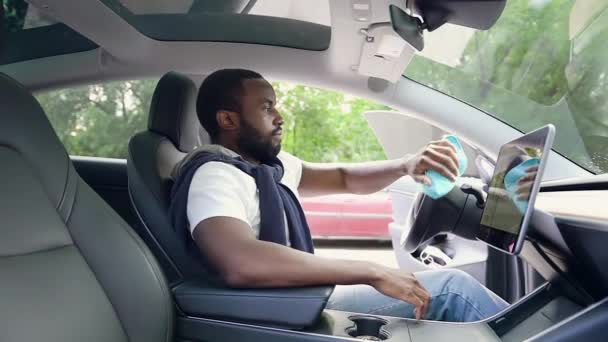Вид збоку красивий юний бородатий чорношкірий чоловік, який сидить всередині власного автомобіля і протирає кермо і екран навігатора ганчіркою з мікрофібри — стокове відео