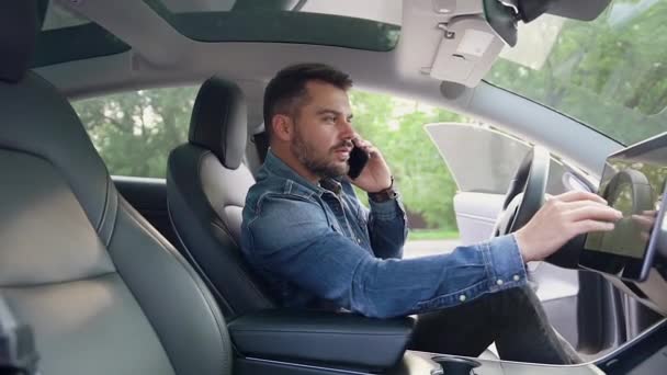 Красивый улыбающийся молодой бородатый мужчина сидит перед штурвалом собственной роскошной машины, разговаривает по телефону и с помощью GPS навигатора — стоковое видео
