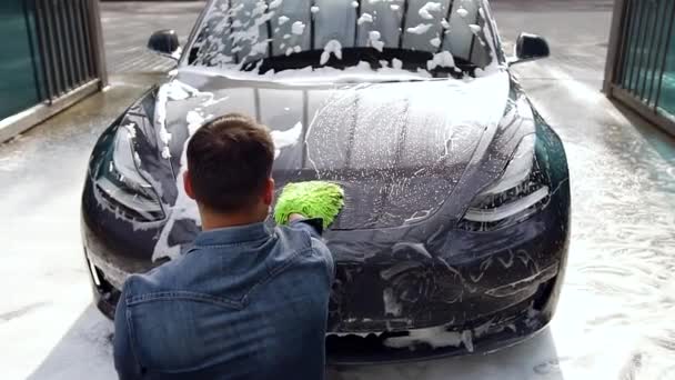 Widok z góry atrakcyjny ciężko pracujący brodaty młody człowiek, który myje swój drogi samochód piankową gąbką w myjni samochodowej — Wideo stockowe