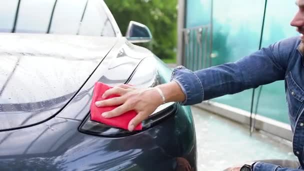 Hezký úsměv mladý vousatý muž otírání reflektory se speciální žíněnkou z mikrovlákna ve venkovním mytí aut