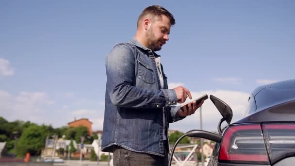 Hombre barbudo agradable que controla el tiempo en el teléfono mientras carga la batería en su lux clase coche eléctrico en la estación de carga especial — Vídeo de stock