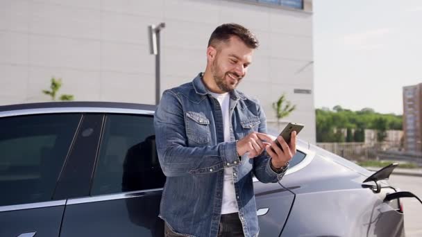 Pozitivní hezký mladý vousatý muž pomocí telefonu při nabíjení jeho luxusní elektromobil v nabíjecí stanici — Stock video