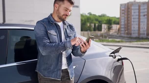 Sympathischer bärtiger Mann, der mit Handy in der Nähe seines luxuriösen Hybridautos steht und den Akku mit einem speziellen Ladegerät in der Ladestation auflädt — Stockvideo